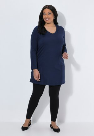 Donna che indossa una tunica blu navy a maniche lunghe, leggings neri e ballerine nere, in piedi su sfondo bianco, sorridente e che guarda di lato.