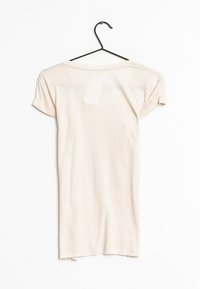 Světle béžové tričko s krátkým rukávem a ohrnutými manžetami, vyrobené z měkké látky a s kulatým výstřihem. Visí na černém kovovém háčku na bílém pozadí.