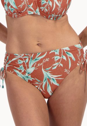 Kobieta nosząca bikini z wysokim stanem na tle pomarańczowego tła, z białym i turkusowym kwiatowym wzorem, posiadające wiązania po bokach na biodrach.