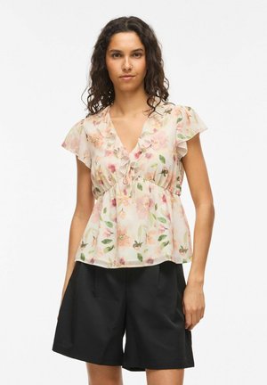 Donna con capelli ricci indossa una blusa floreale con volant e scollo a V e shorts neri a vita alta, in piedi davanti a uno sfondo semplice.