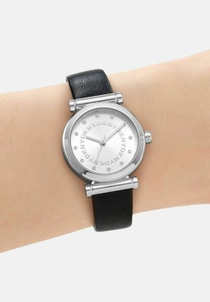 Ronde zilveren horloge met zwarte leren band om de pols, zilveren wijzerplaat met herhaalde "DKNY" tekst en kleine kristallen uurmarkeringen.