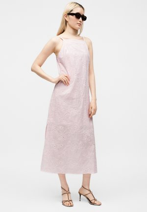 Femme portant une robe midi sans manches rose pâle brodée de fleurs, lunettes de soleil noires et sandales à talons marron à brides, debout, main sur la hanche.
