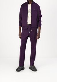 Chándal deportivo morado con chaqueta con cremallera y pantalones con cordón, ambos con un logo discreto. Tejido texturizado, complementado con zapatillas negras.