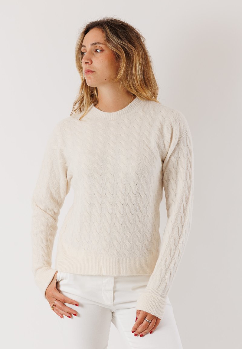 Donna dai capelli lunghi fino alle spalle, che indossa un maglione in maglia di trecce color crema e pantaloni bianchi, che guarda verso sinistra su uno sfondo semplice.