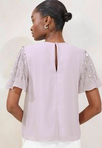Lavender blouse with short, ruffled sleeves adorned with sequins. Features a back keyhole opening and smooth, lightweight fabric texture.
