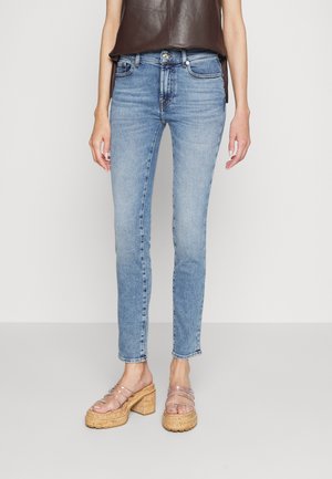 Mujer con vaqueros pitillo azul claro, top sin mangas marrón oscuro y sandalias de plataforma con tiras transparentes sobre un fondo liso y claro.