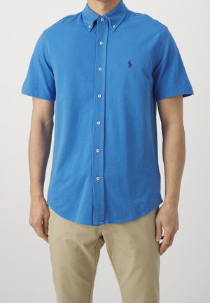 Mies, joka pitää päällään sinistä, lyhythihaista nappipaitaa, jossa on pieni kirjailtu logo rinnassa, ja beiget housut.