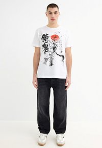 Camiseta de algodón blanca con arte gráfico en negro y rojo, con diseños de inspiración asiática, combinada con pantalones de mezclilla negros y zapatillas blancas.