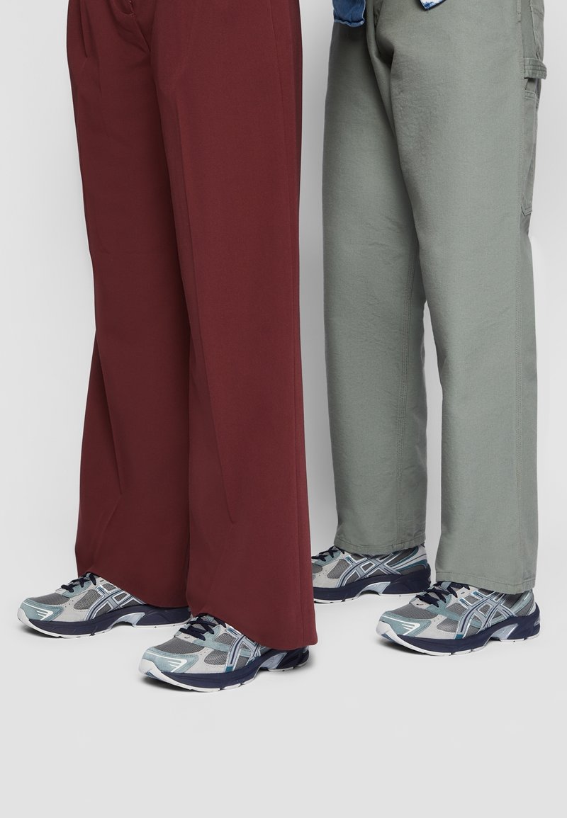 Deux personnes debout côte à côte portant des pantalons larges gris et bordeaux avec des chaussures de course gris clair et bleu sur un fond gris.