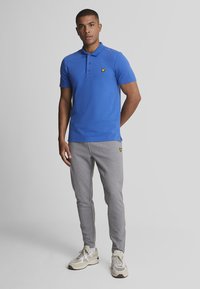 Polo shirt blu in cotone con tre bottoni e logo giallo sul petto, abbinato a pantaloni jogging grigi e scarpe da ginnastica beige.