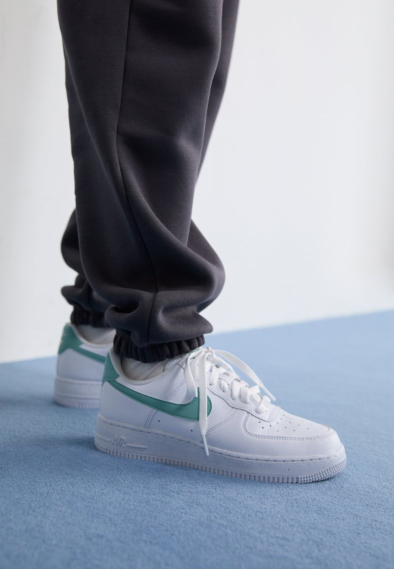 Bijele Nike Air Force 1 tenisice s mint zelenim swooshom, gornjište od teksturirane kože i bijele vezice, u kombinaciji s tamnosivim joggericama s manžetama.