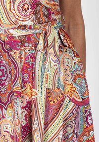 Nærbillede af farverigt paisley-mønsterstof i orange, pink og teal nuancer bundet med en sløjfe på et tøj, der bæres af en person med brun hud.