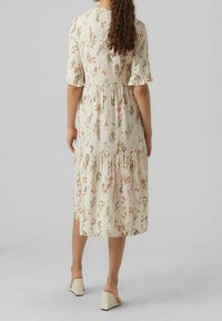 Kwiatowa sukienka do kolan z krótkimi falbaniastymi rękawami, na kremowym tle z zielonymi i różowymi kwiatami, o warstwowej konstrukcji, noszona z sandałami na obcasie bloczkowym.