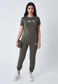 Conjunto en verde oliva que incluye una camiseta ajustada con texto blanco y pantalones jogger con cintura y puños elásticos. Se combina con zapatillas blancas.