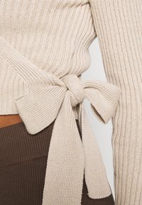 Beżowy sweter z prążkowanego dzianiny z związanym wstążką w talii. Tekstura jest gładka i miękka, z długimi rękawami i dopasowanym fasonem.