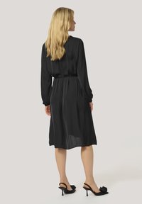 Robe noire à manches longues et taille cintrée, avec une jupe fluide arrivant mi-mollet. Portée avec des mules à talons noirs.