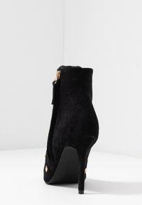 Bottine en daim noir avec une pointe pointue, un talon stiletto haut et un détail de fermeture éclair doré. Surface texturée avec des accents découpés.