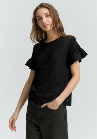 Sort struktureret kortærmet top med flæseærmer; afslappet pasform; båret med mørke denimbukser. Har en rund halsudskæring og sideaccentuer.