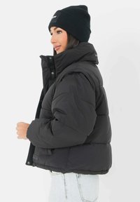 Zwarte pufferjack met hoge kraag, gewatteerd ontwerp en knoopsluiting. Gepaard met een zwarte gebreide beanie. Lichtgekleurde spijkerbroek.