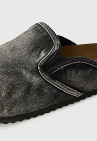 Zwarte denim instapschoen met textuur op de bovenkant, contrasterende stiksels langs de naad en een flexibele rubberen zool. Het interieur heeft een bruine voering.