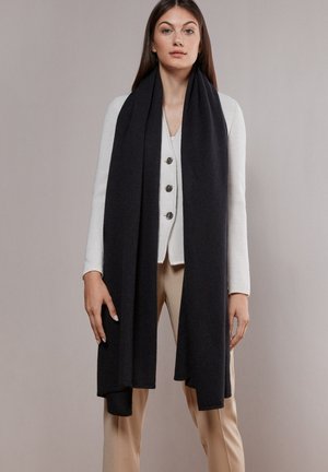 Jonge vrouw met een lange zwarte sjaal over een witte gebreide cardigan met knopen en beige broek, staand tegen een effen grijze achtergrond.