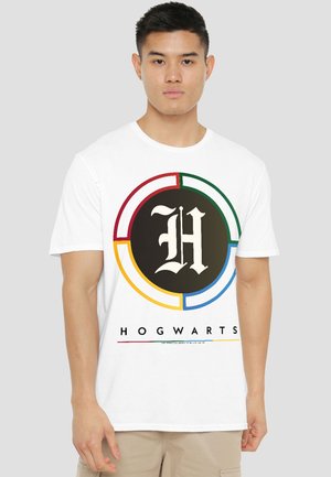Young man wearing a white T-shirt with a multicolored circular Hogwarts emblem and "Hogwarts" text below on a plain background.