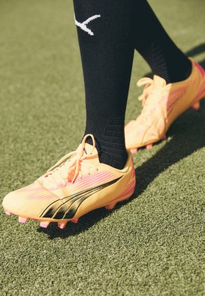 Ryškiai geltoni futbolo batai su juodais akcentais ir rožiniais studais, turintys elegantišką dizainą ir tekstūruotą paviršių. Nešioti su juodais kelio ilgio raišteliais.