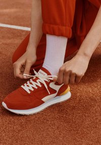 Zapatillas rojas con detalles en blanco, materiales de ante y malla, con suela plana, punta redondeada y logo en la lengüeta. Se están atando los cordones.