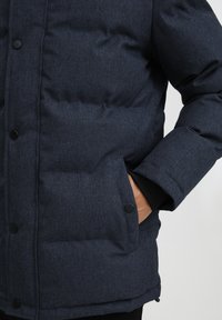 Marinblå pufferjacka med texturerad finish, som har knäppning med knappar, sidofickor och elastiska ärmslut. Varm och isolerad design.