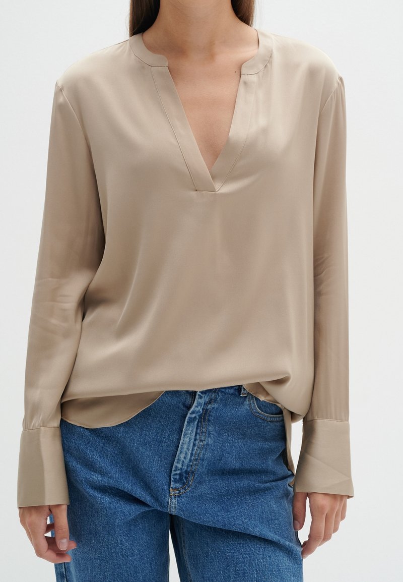 Femme portant un chemisier beige à manches longues avec un col en V et un jean bleu, debout devant un fond uni clair.
