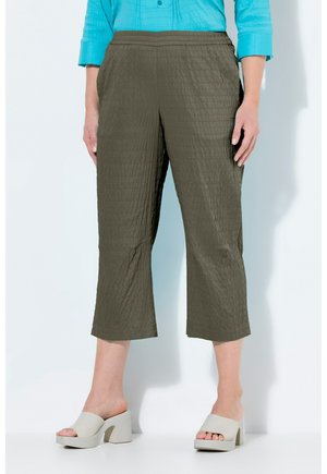 Modelka w oliwkowozielonych, teksturowanych spodniach o kroju cropped oraz jasnoniebieskiej bluzce z długim rękawem, w zestawieniu z beżowymi sandałami na wysokim obcasie z paseczkiem z tyłu.