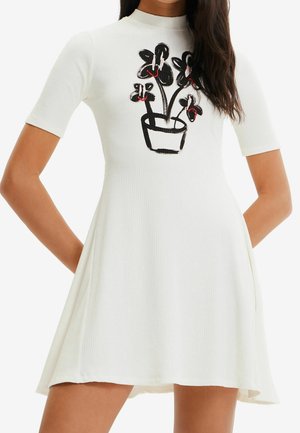 Hvid kortærmet ribbet kjole med sort og rød abstrakt blomsterprint på brystet, båret af en person med langt mørkt hår.