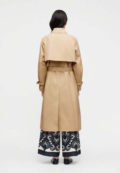 Γυναίκα με μακριά σκουρόχρωμα μαλλιά, φορώντας ένα μπεζ παλτό τύπου τραντσόμπουκα πάνω από φαρδιά, μαύρα και άσπρα παντελόνια με σχέδιο, στέκεται με την πλάτη γυρισμένη σε μονόχρωμο φόντο.