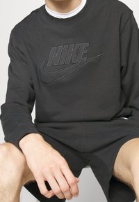 En person sitter i en svart Nike-tröja och matchande shorts, med Nike-logotypen på bröstet.