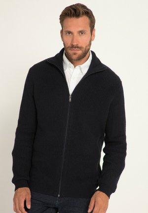 Maglioncino zip con cappuccio di colore blu navy, con una texture a coste, colletto alto e maniche lunghe. Indossato sopra una camicia bianca con bottoni, design attillato.