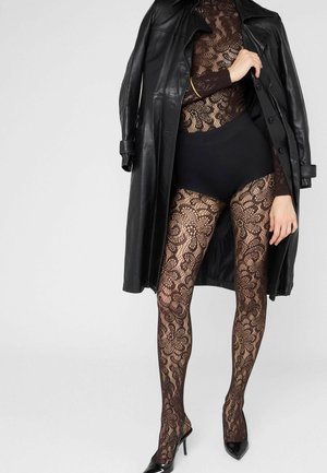 Personne portant un body en dentelle noire, des collants en dentelle à motifs, des talons hauts noirs et un trench-coat en cuir noir drapé sur les épaules.