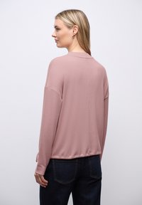 Langärmliges Oberteil aus weichem, pinkfarbenem Stoff mit lockerer Passform, Rundhalsausschnitt und leicht cropped Saum, kombiniert mit dunkelblauen Jeans.