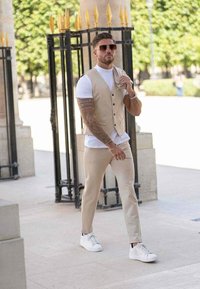 Gilet beige sur un t-shirt blanc, associé à un pantalon beige et des baskets blanches. Lunettes de soleil, tatouages et détail d'une chemise décontractée repliée.