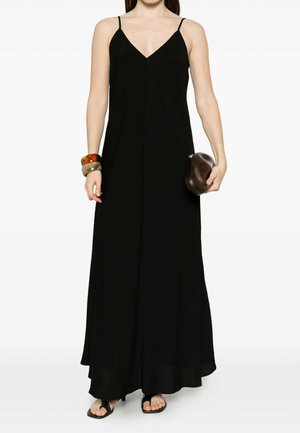 Donna che indossa un lungo vestito nero senza maniche con sottili spalline, che tiene in mano una pochette marrone e indossa due bracciali spessi a un polso.