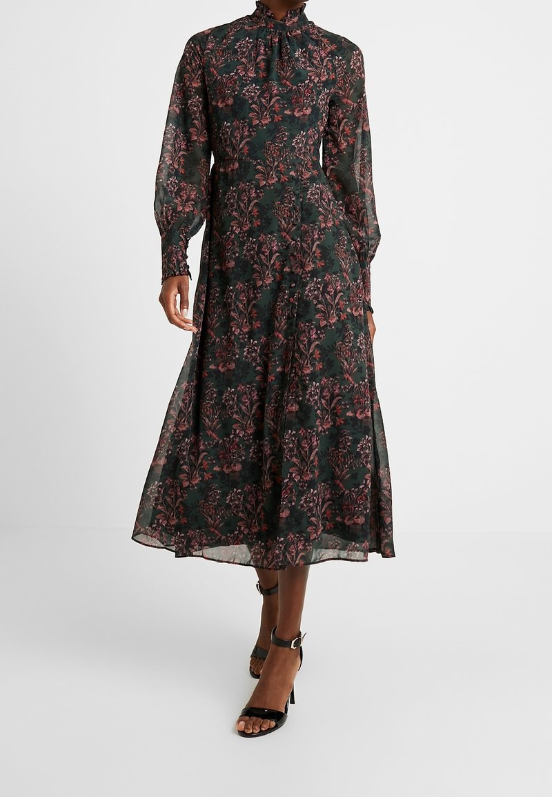 Pitkä, tummanvihreä mekko, jossa on pinkkejä ja punaisia kukkakuoseja. Sisältää korkean kauluksen, pitkät hihat ja hulmuavan helman. Mustat remmi-korkokengät.