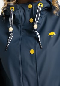 Marinblå regnjacka i vattentätt material med gula knappar och detaljer, justerbara snörningar med träperlor och en paraplylogotyp.