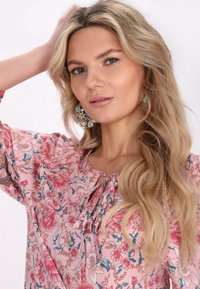 Bluză florală roz cu mâneci bufante, prezentând un detaliu cu decolteu în formă de cheie și modele intricate. Cercei mari, decorative completează aspectul.