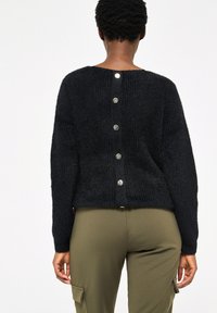 Černý pletený svetr s nízkým výstřihem na zádech a pěti ozdobnými stříbrnými knoflíky. Lehce zkrácený střih s dlouhými rukávy a měkkou texturou.