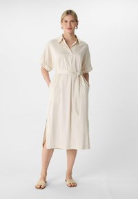 Beiges Hemdkleid mit Kragen und kurzen, hochgerollten Ärmeln, ausgestattet mit einem Bindegürtel in der Taille, Seitentaschen und seitlichen Schlitzen. Getragen mit neutralen Sandalen.