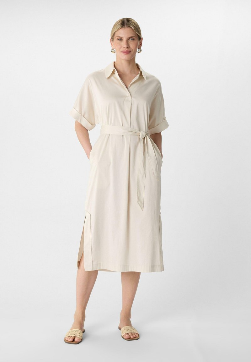 Beiges Hemdkleid mit Kragen und kurzen, hochgerollten Ärmeln, ausgestattet mit einem Bindegürtel in der Taille, Seitentaschen und seitlichen Schlitzen. Getragen mit neutralen Sandalen.