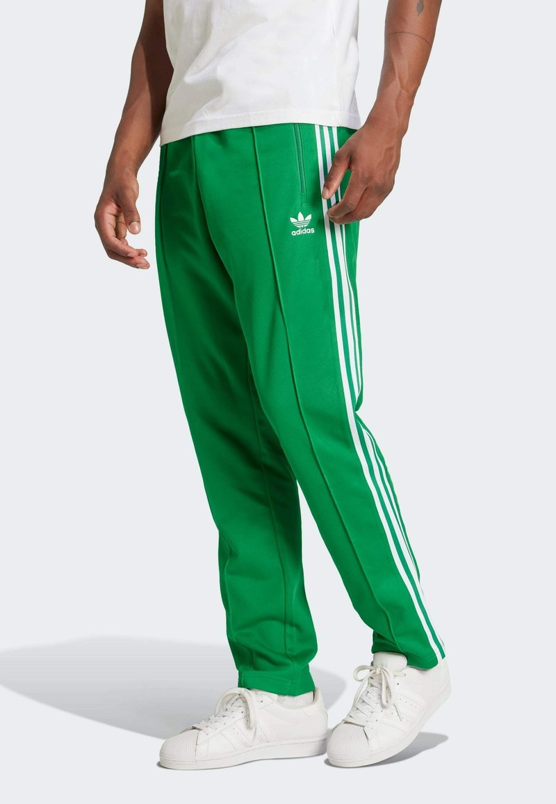 Pantalones deportivos verdes con franjas laterales blancas y logo. Textura suave y diseño ajustado, combinados con zapatillas blancas.