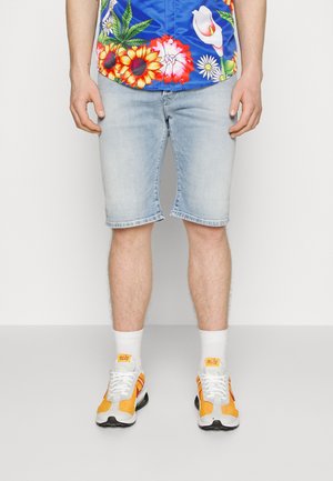 Blaue Bluse mit kurzen Ärmeln und lebhaften Blumenmustern, kombiniert mit hellblauen Denim-Shorts und mehrfarbigen Turnschuhen.