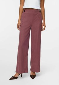 Vidjejeans i ett mjukt mauve-tyg, med hög midja och framknappar samt en slät textur. Matchat med spetsiga bruna klackar.