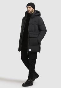 Black puffer jacket with a hood and multiple pockets, featuring a zip front and logo detail. Worn with black pants and sturdy boots.