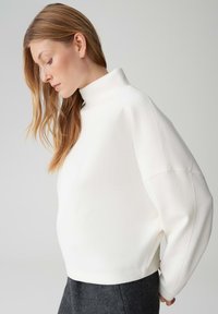 Weißes, kurz geschnittenes Sweatshirt mit hohem Kragen, weicher Textur und überschnittenen Schultern, kombiniert mit grauen, maßgeschneiderten Hosen. Einfaches und modernes Design.